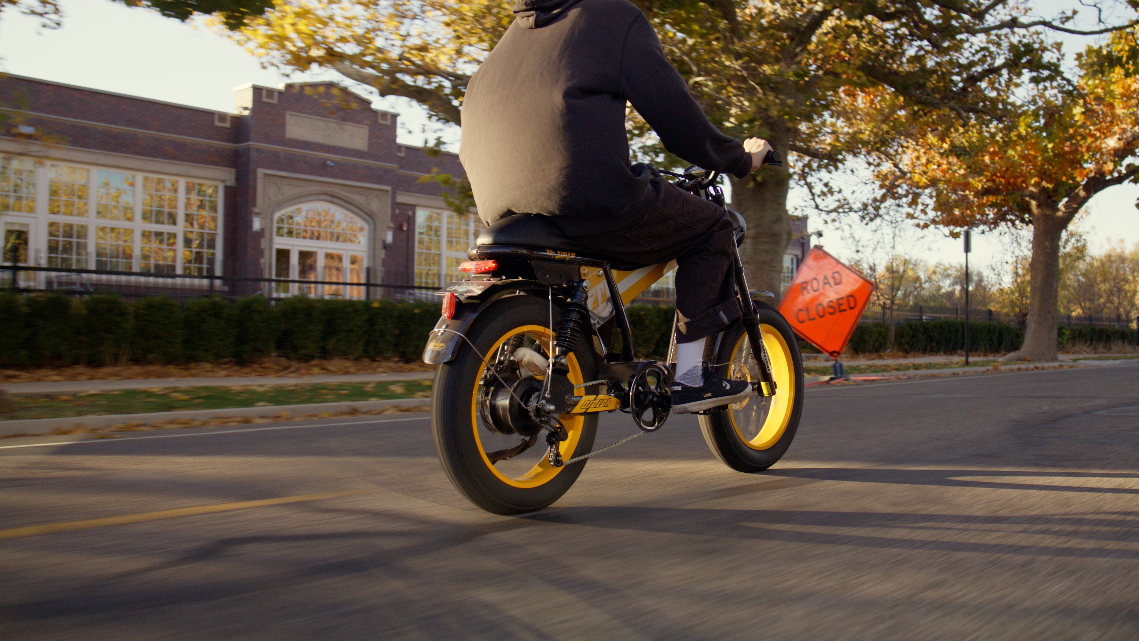 Rider cruising on a fat tire electric bike with yellow rims on a city street, passing a road closed sign in autumn