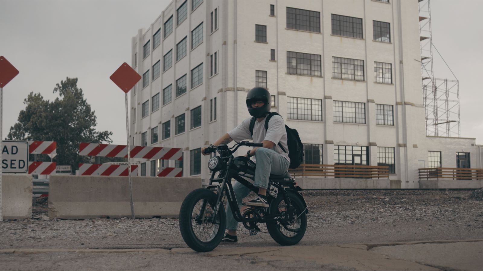 Rider wearing a helmet and backpack on a Qiolor Tiger RE electric bike, paused on an urban street with industrial buildings and construction barriers in the background.